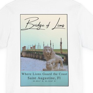 May include: White t-shirt with a graphic on the back featuring the Bridge of Lions in Saint Augustine, Florida. The graphic includes a bridge scene with a lion statue, text that reads 'Bridge of Lions,' and 'Where Lions Guard the Coast Saint Augustine, Fl' with coordinates.