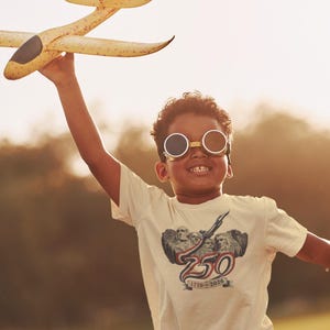 May include: A cream-colored short-sleeve t-shirt featuring a graphic print of Mount Rushmore with the text 250 and 1776-2020. The shirt is worn with round, dark-tinted goggles with a metallic frame, and a yellow foam toy airplane is held aloft.