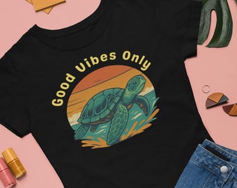 May include: Black t-shirt with a graphic of a sea turtle in front of a sunset, with the words "Good Vibes Only" in yellow. The shirt is paired with a denim skirt and sandals on a pink background.
