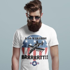 Puede incluir: Camiseta blanca con un gr&aacute;fico del A-10 Warthog. El dise&ntilde;o incluye el texto "A-10 WARTHOG" y "BRRRRRTT!!" encima y debajo del avi&oacute;n, sobre un c&iacute;rculo rojo, blanco y azul con estrellas y rayas, y un emblema de la Fuerza A&eacute;rea de EE. UU.