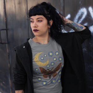 May include: Gray t-shirt featuring a moth design with a crescent moon and starry night sky. The moth is brown with orange accents. The shirt is paired with a plaid skirt and black jacket.