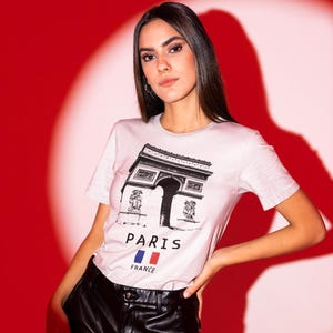 Puede incluir: Camiseta rosa claro con un dibujo en blanco y negro del Arco del Triunfo. Las palabras "PARIS" y "FRANCE" est&aacute;n impresas debajo, junto con la bandera francesa. La persona lleva pantalones de cuero negro.