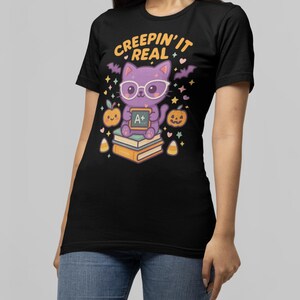 May include: Black t-shirt with a Halloween-themed graphic. The design features a purple cat wearing glasses and holding an A+ sign, with the text "CREEPIN' IT REAL". The cat is sitting on books, with pumpkins and bats.