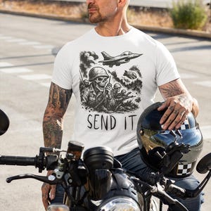 May include: White t-shirt with a black graphic of a skull soldier, fighter jets, and the text "SEND IT." The shirt is worn by a person sitting on a black motorcycle, holding a black helmet. The motorcycle has a round headlight.