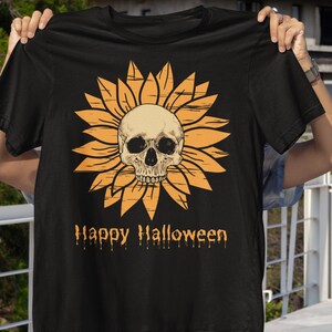 May include: Black t-shirt featuring a sunflower design with a skull in the center. The sunflower is orange, and the skull is off-white. The words "Happy Halloween" are printed below the design in orange.
