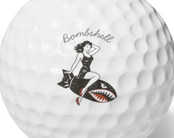 May include: A white golf ball with a dimpled surface featuring a graphic of a person in a black dress riding a black bomb with red accents and sharp teeth. The word Bombshell is printed in a cursive script above the graphic illustration.