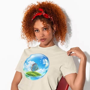 May include: A beige short-sleeved t-shirt featuring a graphic print of a blue fish swimming inside a transparent bubble that contains a small grassy island under a bright blue sky with white clouds and rising air bubbles.