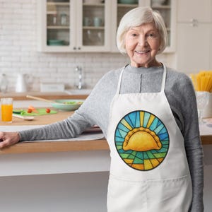 May include: White apron with a colorful, circular design featuring a yellow empanada-like pastry. The design is framed by blue and yellow rays, resembling stained glass. The apron has a pocket and is worn by a person in a kitchen setting.