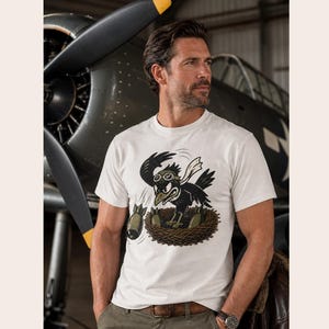Puede incluir: Una camiseta blanca de manga corta con una ilustraci&oacute;n gr&aacute;fica de un p&aacute;jaro negro con gafas de aviador, posado en un nido y lanzando bombas de color verde oliva. El p&aacute;jaro lleva una bufanda blanca y tiene una expresi&oacute;n agresiva de dibujos.