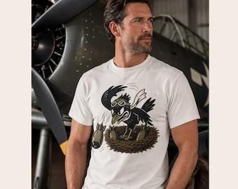 May include: A white short-sleeved t-shirt featuring a graphic illustration of a black bird wearing aviator goggles, perched in a nest and dropping olive green bombs. The bird has a white scarf and an aggressive expression, rendered in a cartoon style.