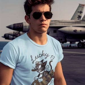 May include: A light blue t-shirt featuring a graphic of a black cat wearing goggles and smoking a cigar while riding a brown bomb. The text Lucky 13 is printed above the cat in a cursive font. The shirt has short sleeves and a crew neckline.