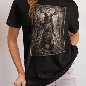 May include: A black short-sleeved t-shirt featuring a rectangular graphic print of a skeletal creature with large antlers standing in a dark forest under a full moon. The illustration is rendered in shades of brown, black, and white.