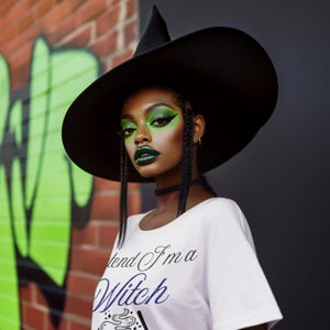 May include: A person wearing a black witch hat and a white t-shirt with the text "I pretend I'm a Witch". The person has green eye makeup and dark green lipstick. The background includes a brick wall and green graffiti.