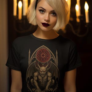 May include: Black t-shirt featuring a gothic gargoyle design with a red stained-glass window. The gargoyle has bat-like wings and is set against a dark background. The design is detailed and artistic.