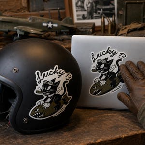 May include: A matte black motorcycle helmet and a silver laptop both featuring a sticker of a black cat wearing goggles and a scarf while riding an olive green bomb. The sticker includes the text Lucky 13 in a white cursive script font.
