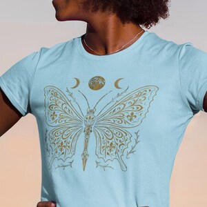May include: Light blue t-shirt featuring a gold butterfly design. The butterfly has intricate patterns and is centered on the shirt. Above the butterfly are a crescent moon and a circle. The shirt is a crew neck and short-sleeved.