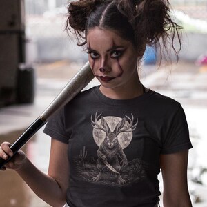 K&ouml;nnte beinhalten: Ein dunkelgraues Kurzarm-T-Shirt mit einem Grafikdruck eines Jackalopen, der ein Messer in einer W&uuml;stenlandschaft vor einem Vollmond h&auml;lt. Dazu werden wei&szlig;e Jeansshorts mit ausgefranstem Saum und schwarze Overknee-Socken getragen.