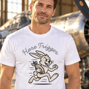 Puede incluir: Una camiseta blanca de manga corta con un gr&aacute;fico de un conejo de dibujos animados corriendo mientras sostiene una bomba. Sobre el conejo, el texto "Hare Trigger" est&aacute; impreso en una fuente cursiva oscura. El dise&ntilde;o usa tonos beige, marr&oacute;n y negro.