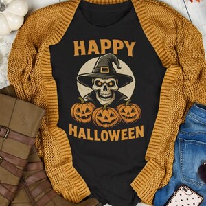 May include: A flat lay featuring a black t-shirt with a skull wearing a witch hat and the words "Happy Halloween". The shirt is paired with a mustard yellow cardigan, blue jeans, and brown boots. Decorative pumpkins and fall leaves surround the items.