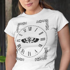 May include: White t-shirt featuring a black clock design with Roman numerals and a moth graphic at the center. The clock is framed by decorative black flourishes. The model is wearing gray pants and standing in front of a textured background.