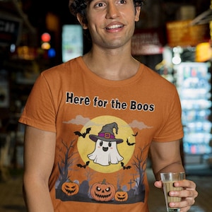 May include: Orange t-shirt with the text "Here for the Boos" and a Halloween graphic featuring a ghost, bats, and pumpkins. The person is wearing a brown hat and khaki pants.