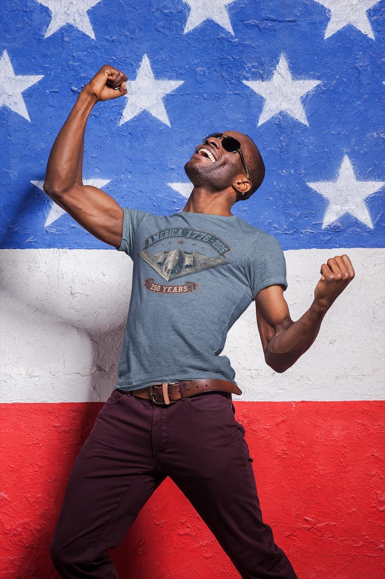 Puede incluir: Una camiseta azul jaspeado de manga corta con un gr&aacute;fico de un bombardero furtivo. El texto dice "AMERICA 1776-2026" y "250 YEARS" en un dise&ntilde;o de pancarta. La camiseta se combina con pantalones de color granate oscuro y un cintur&oacute;n de cuero marr&oacute;n.