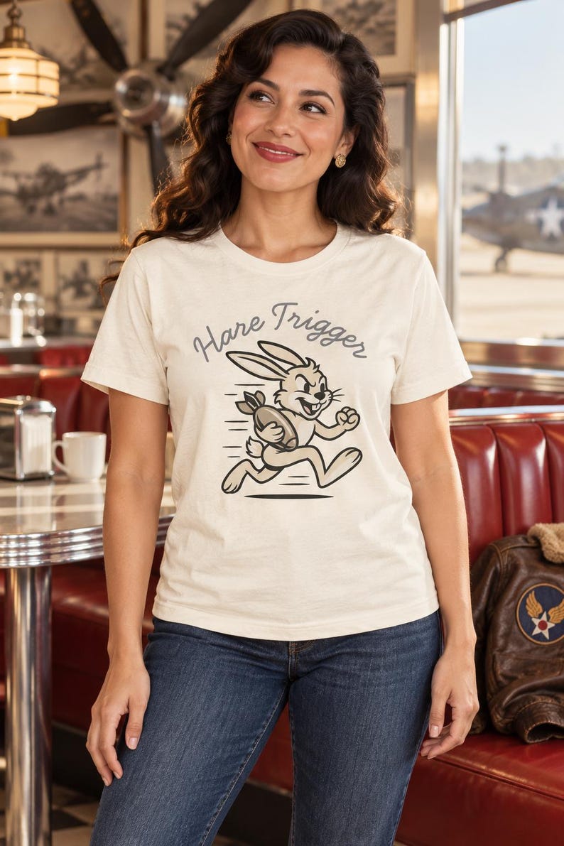 Puede incluir: Una camiseta de manga corta color crema con un gr&aacute;fico de un conejo de dibujos animados corriendo mientras sostiene una bomba. El texto "Hare Trigger" est&aacute; impreso en una fuente gris oscuro arqueada sobre la ilustraci&oacute;n. Cuello redondo.