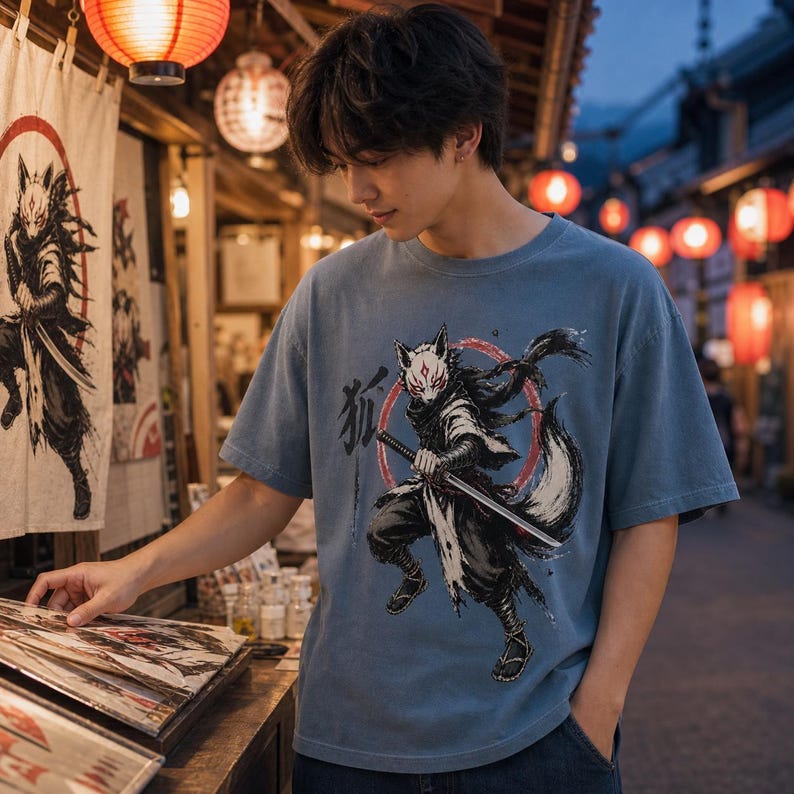 May include: A dusty blue short-sleeved t-shirt featuring a graphic print of a fox-masked warrior holding a katana sword. The illustration includes a red circular background element and black Japanese kanji characters on the left side of the design.