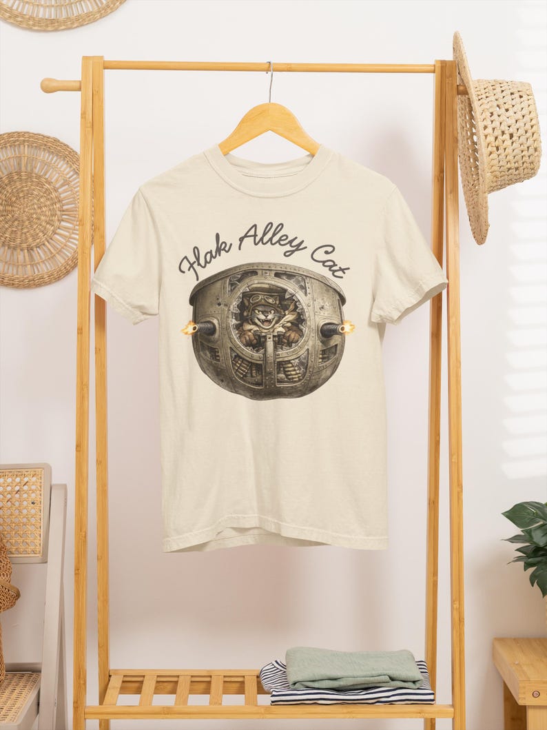 以下が含まれることがあります： クリーム色の半袖Tシャツ。飛行帽とゴーグルを着用した猫が金属製の砲塔の中にいるグラフィックが描かれています。イラストの上部には、暗い筆記体フォントで「Flak Alley Cat」という文字がプリントされています。