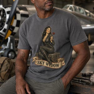 May include: A charcoal gray t-shirt featuring a graphic of a person in a dark flight suit sitting on a crate, holding a red rose. Below the figure is a tan banner with the text "SKY WYDOW" in black letters, accented with two small red stars.