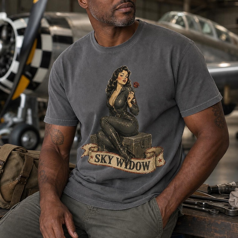 May include: A charcoal gray t-shirt featuring a graphic of a person in a dark flight suit sitting on a crate, holding a red rose. Below the figure is a tan banner with the text "SKY WYDOW" in black letters, accented with two small red stars.