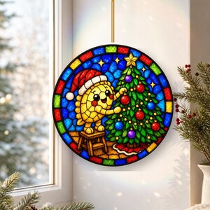 May include: A circular stained glass suncatcher featuring a yellow character wearing a red Santa hat, standing on a small stool to decorate a green Christmas tree with red and purple ornaments, all framed by a colourful mosaic border.