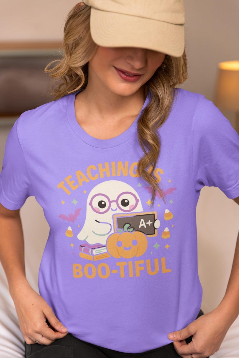 May include: A lilac t-shirt with a Halloween-themed graphic. The design features a ghost wearing glasses, a pumpkin, and the words "TEACHING'S BOO-TIFUL" in orange and yellow. The shirt also includes bats, candy corn, and an A+ grade.