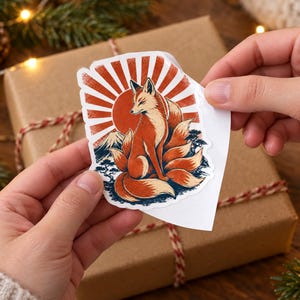 May include: A die-cut sticker featuring a multi-tailed fox sitting in front of a large red sun, with a mountain silhouette and dark blue landscape elements at the base. The design uses a vibrant color palette of orange, red, and deep blue tones.