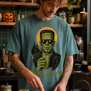 Może przedstawiać: Turkusowy t-shirt z okrągłym dekoltem i grafiką przedstawiającą zielonego potwora w okularach przeciwsłonecznych, pijącego zielony nap&oacute;j. Wz&oacute;r zawiera ż&oacute;łte koło w tle, sylwetkę ciemnego zamku oraz ż&oacute;łty napis Matcha Monster.