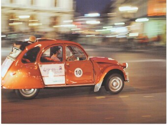 May include: An orange vintage car, with the number 10 on the door, is driving on a city street at night. The car has a white door and a red roof. The background is blurred with city lights.