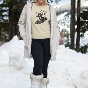 May include: A beige sweatshirt with a graphic of a black cat riding a bomb, with the text "Lucky 13". The person is wearing a brown beanie, a light-colored coat, black jeans, and white snow boots with fur trim. The background is a snowy landscape.