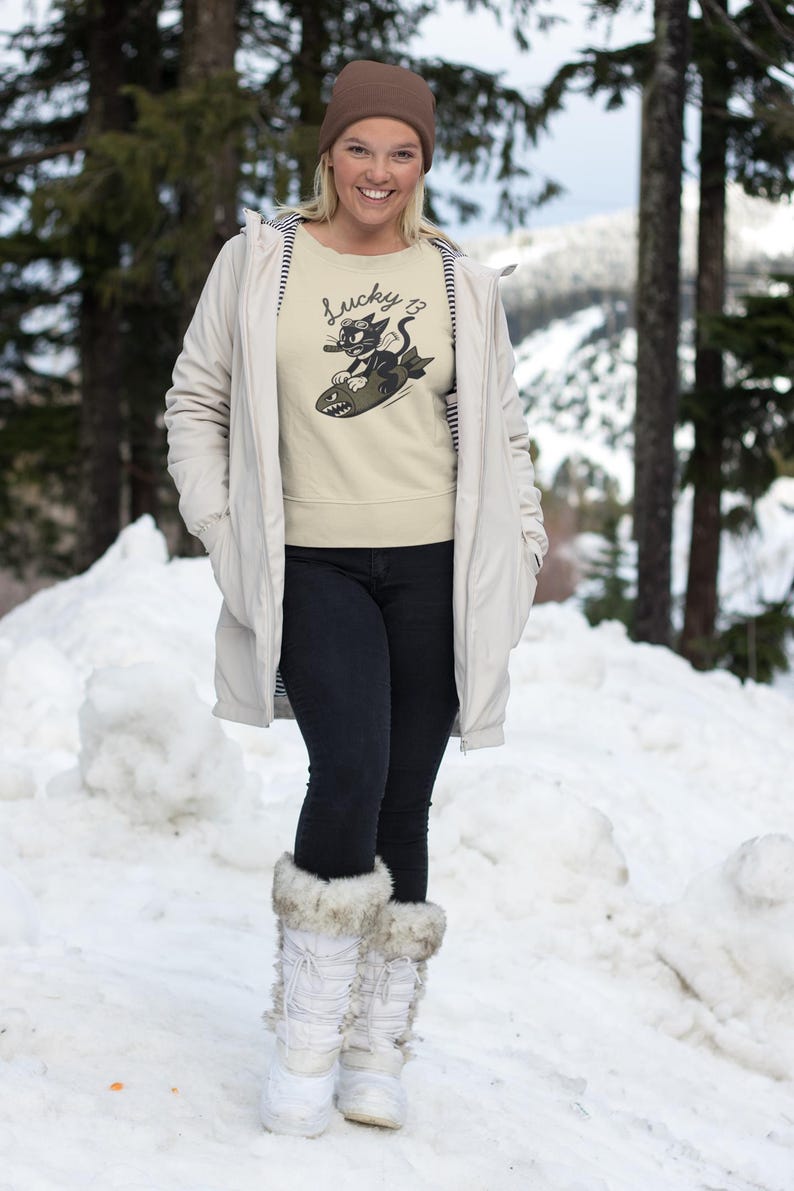 May include: A beige sweatshirt with a graphic of a black cat riding a bomb, with the text "Lucky 13". The person is wearing a brown beanie, a light-colored coat, black jeans, and white snow boots with fur trim. The background is a snowy landscape.