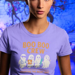 May include: A lilac-colored t-shirt with the text "BOO BOO CREW" in orange. Below the text are four cartoon ghosts dressed as medical professionals. The phrase "HAUNTING THE NIGHT SHIFT" is printed below the ghosts.
