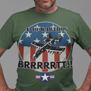 May include: Green t-shirt featuring a graphic of an A-10 Warthog aircraft. The design includes the text "A-10 WARTHOG" and "BRRRRTT!!" in a circular emblem with stars and stripes, and a blue star with red and white stripes.