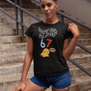 Puede incluir: Camiseta negra con el texto "DON'T ASK I'M JUST 67" en blanco y rojo. Un pollito de dibujos animados con un bocadillo "meh" est&aacute; debajo del texto. La modelo lleva pantalones cortos vaqueros.