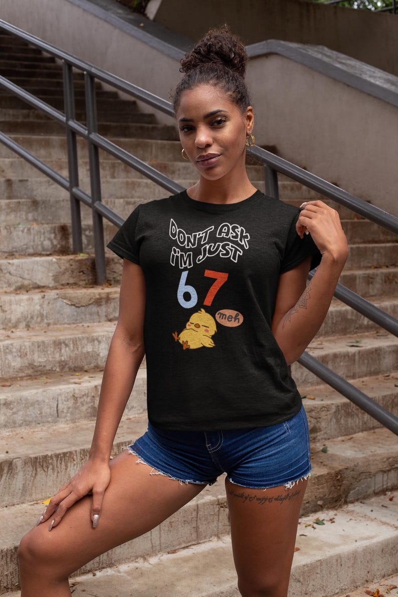 Puede incluir: Camiseta negra con el texto "DON'T ASK I'M JUST 67" en blanco y rojo. Un pollito de dibujos animados con un bocadillo "meh" est&aacute; debajo del texto. La modelo lleva pantalones cortos vaqueros.