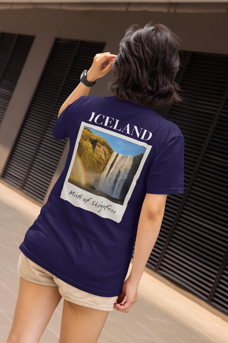 May include: Navy blue t-shirt with a polaroid-style graphic of a waterfall and rainbow, with the word "ICELAND" above and "Mists of Sk&oacute;gafoss" below. The shirt is a short-sleeved crew neck.