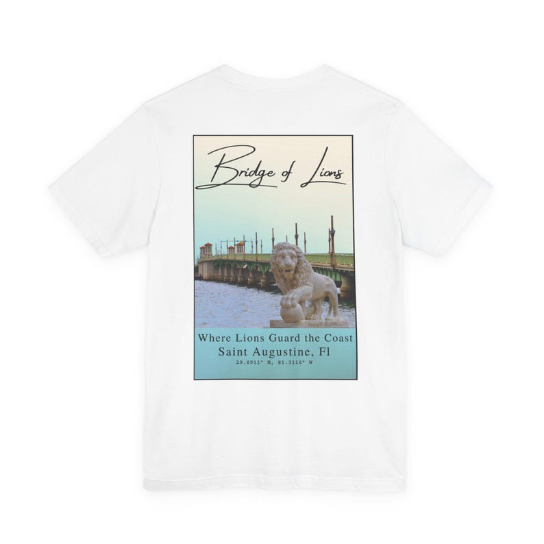 May include: White t-shirt with a graphic on the back featuring the Bridge of Lions in Saint Augustine, Florida. The graphic includes a bridge scene with a lion statue, text that reads 'Bridge of Lions,' and 'Where Lions Guard the Coast Saint Augustine, Fl' with coordinates.