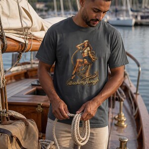 May include: A charcoal gray short-sleeve t-shirt featuring a graphic print of a pin-up sailor sitting on a large metal anchor with a rope coiled around it. The text "Anchors Aweigh" is printed in a gold script font beneath the anchor illustration.