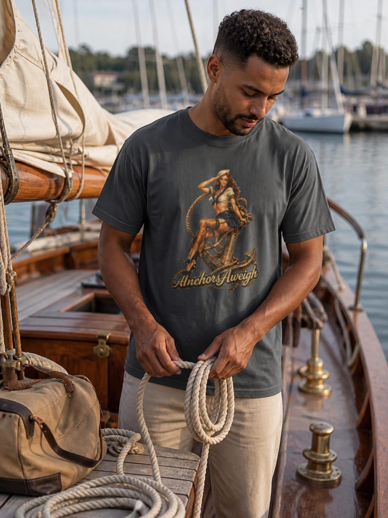 May include: A charcoal gray short-sleeve t-shirt featuring a graphic print of a pin-up sailor sitting on a large metal anchor with a rope coiled around it. The text "Anchors Aweigh" is printed in a gold script font beneath the anchor illustration.