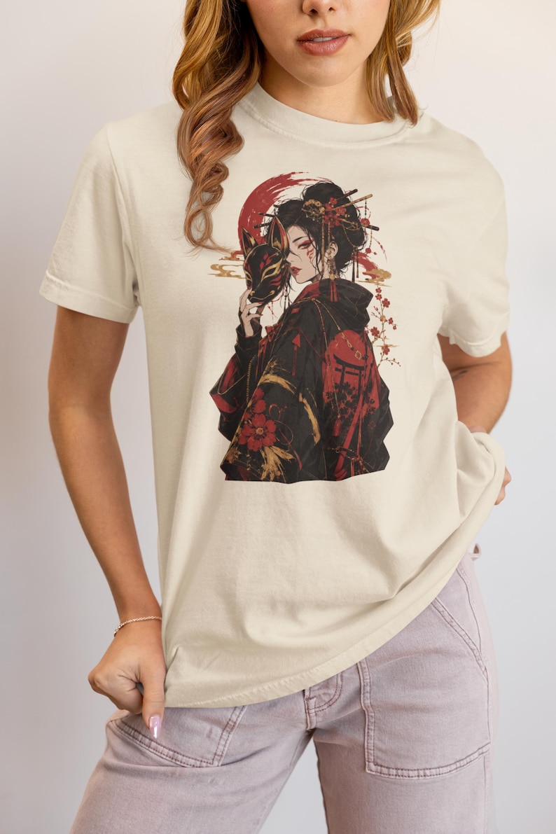 May include: A cream-colored short-sleeve t-shirt featuring a graphic print of a person in a black kimono holding a fox mask. The design includes red floral accents, a red circular background element, and gold details, set against a neutral background.