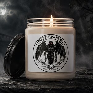 May include: A clear glass jar candle with a lit wick and a black lid. The white label features a circular black graphic of a winged creature with red eyes, text reading POINT PLEASANT WV 1966, and coordinates 38.8443 N, 82.1385 W.