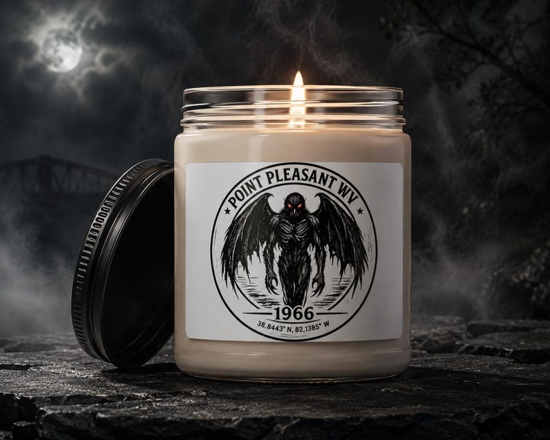 May include: A clear glass jar candle with a lit wick and a black lid. The white label features a circular black graphic of a winged creature with red eyes, text reading POINT PLEASANT WV 1966, and coordinates 38.8443 N, 82.1385 W.
