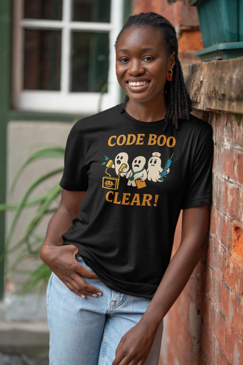 May include: Black t-shirt with the text "CODE BOO CLEAR!" in orange. The graphic features cartoon ghosts with medical equipment. The person is wearing light blue jeans and is leaning against a brick wall.